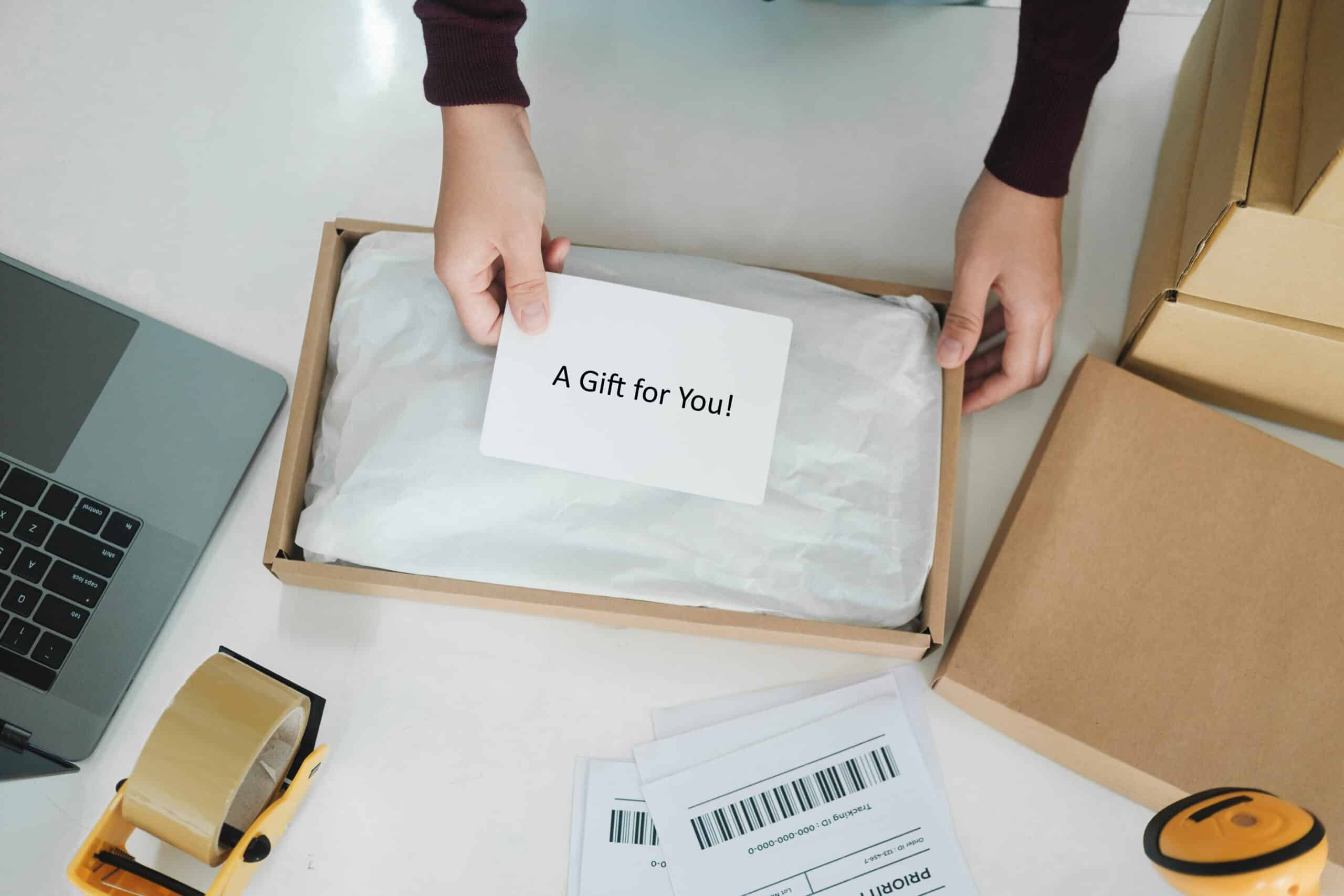  A person is packaging a gift in a brown cardboard box with white paper. They are holding a card that says "A Gift for You!" The scene includes a laptop, packing tape, shipping labels, and other cardboard boxes on a white table.