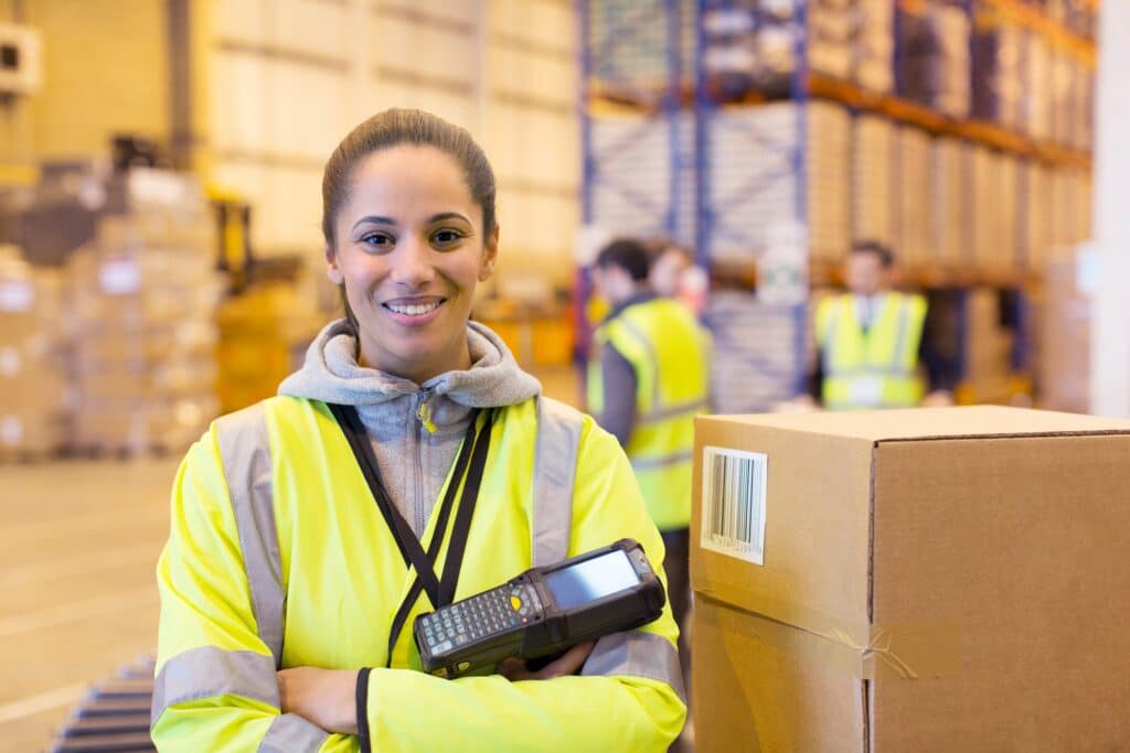 A smiling warehouse worker wearing a high-visibility yellow jacket stands in a large warehouse with shelves stocked with boxes. She holds a barcode scanner